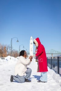 A romantic surprise proposal in the snow at Montréal's Old Port with the white Clock Tower in the background. One person is on one knee while the other reacts with surprise.