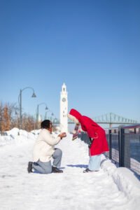 A romantic surprise proposal in the snow at Montréal's Old Port with the white Clock Tower in the background. One person is on one knee while the other reacts with surprise.