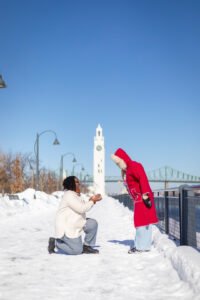 A romantic surprise proposal in the snow at Montréal's Old Port with the white Clock Tower in the background. One person is on one knee while the other reacts with surprise.