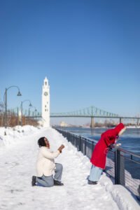 A romantic surprise proposal in the snow at Montréal's Old Port with the white Clock Tower in the background. One person is on one knee while the other reacts with surprise.