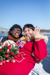 A romantic surprise proposal in the snow at Montréal's Old Port with the white Clock Tower in the background. One person is on one knee while the other reacts with surprise.