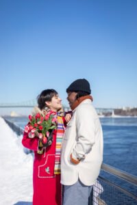 A romantic surprise proposal in the snow at Montréal's Old Port with the white Clock Tower in the background. One person is on one knee while the other reacts with surprise.