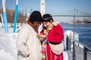 A romantic surprise proposal in the snow at Montréal's Old Port with the white Clock Tower in the background. One person is on one knee while the other reacts with surprise.
