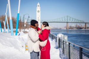 A romantic surprise proposal in the snow at Montréal's Old Port with the white Clock Tower in the background. One person is on one knee while the other reacts with surprise.