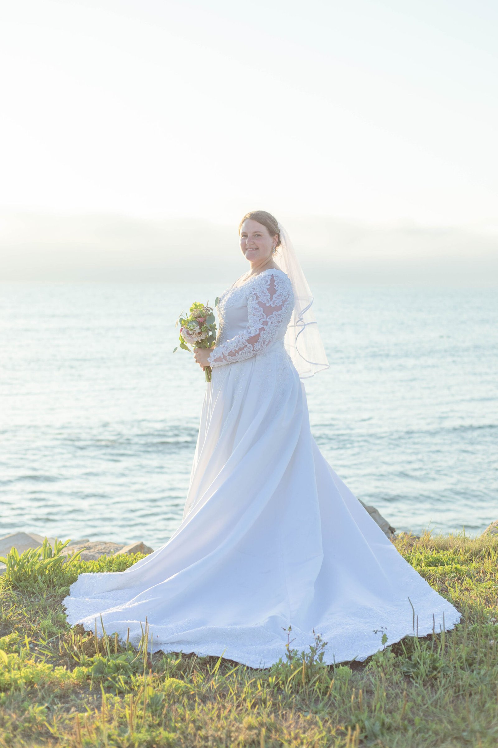 A bride in a white wedding dress standing in a scenic coastal field during a Nova Scotia elopement.