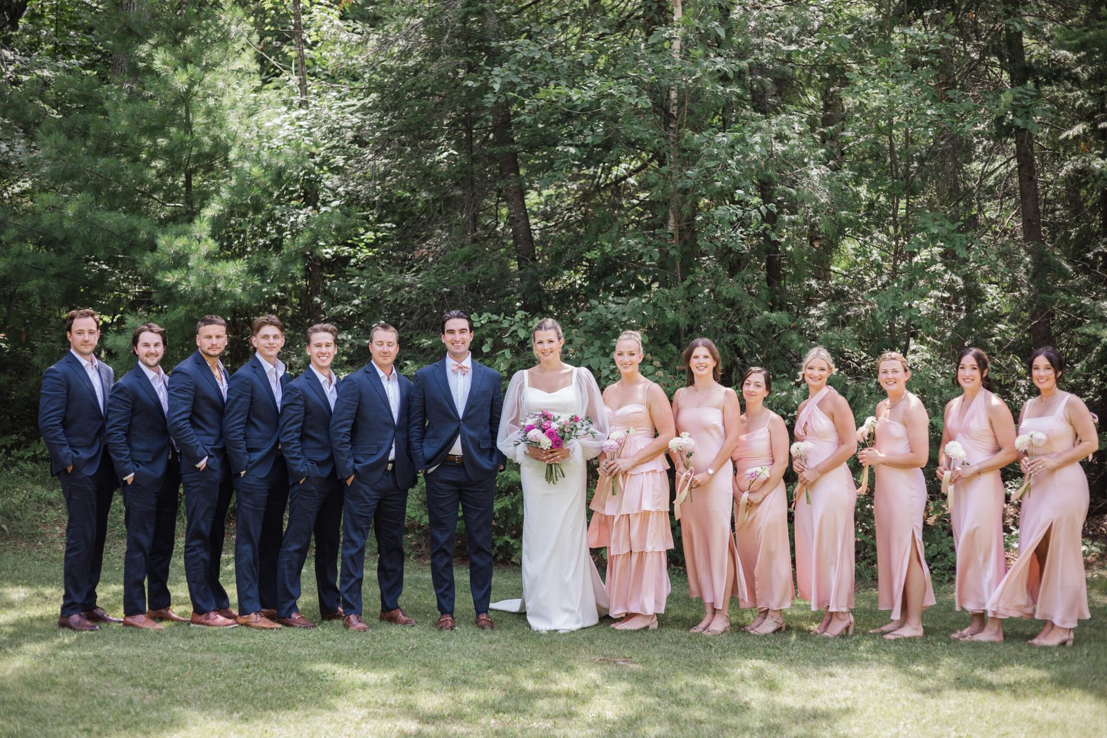 Wedding photography: full wedding party outdoors in a forest clearing, groomsmen in navy suits and bridesmaids in pink dresses