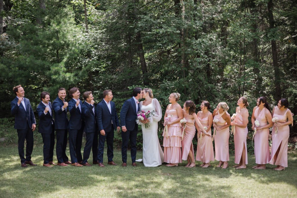 Wedding photography: candid wedding party moment, couple kissing in the centre while the wedding party cheers and laughs around them